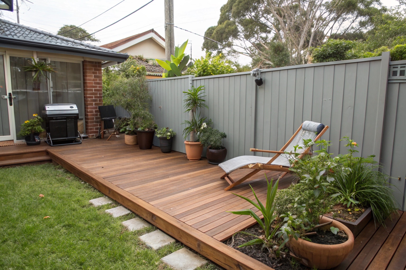 mall cozy timber deck on a compact backyard, spotted gum boards, potted plants along the edge, single lounge chair, small Weber BBQ, fence visible